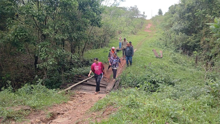 Grupo de seis personas camina por un sendero en área rural, con vegetación densa y un puente de madera sobre un arroyo.