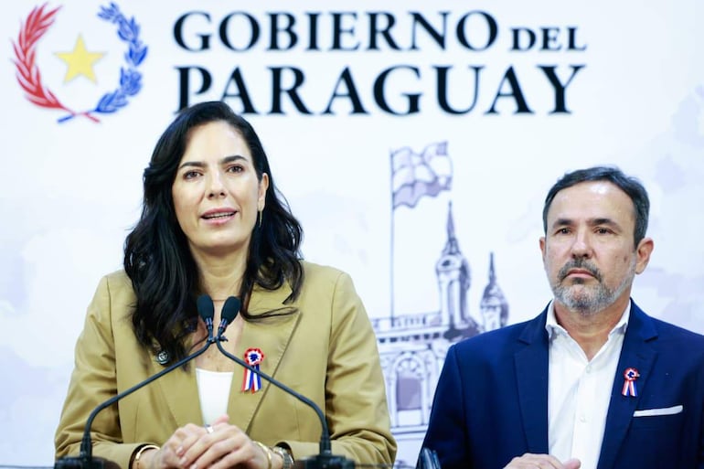Claudia Centurión in a beige jacket in front of a microphone and Emiliano Fernández in a dark blazer next to her, in an official environment.