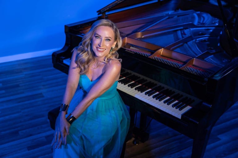 Mujer con cabello rubio y vestido azul claro sonríe junto a un piano brillante en un entorno interior azul.