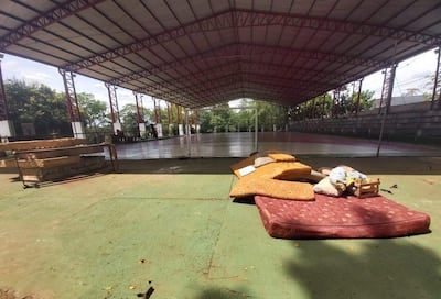 Instalación deportiva vacía con colchones y almohadas en el suelo, techado grande, graderías y árboles visibles en el fondo.