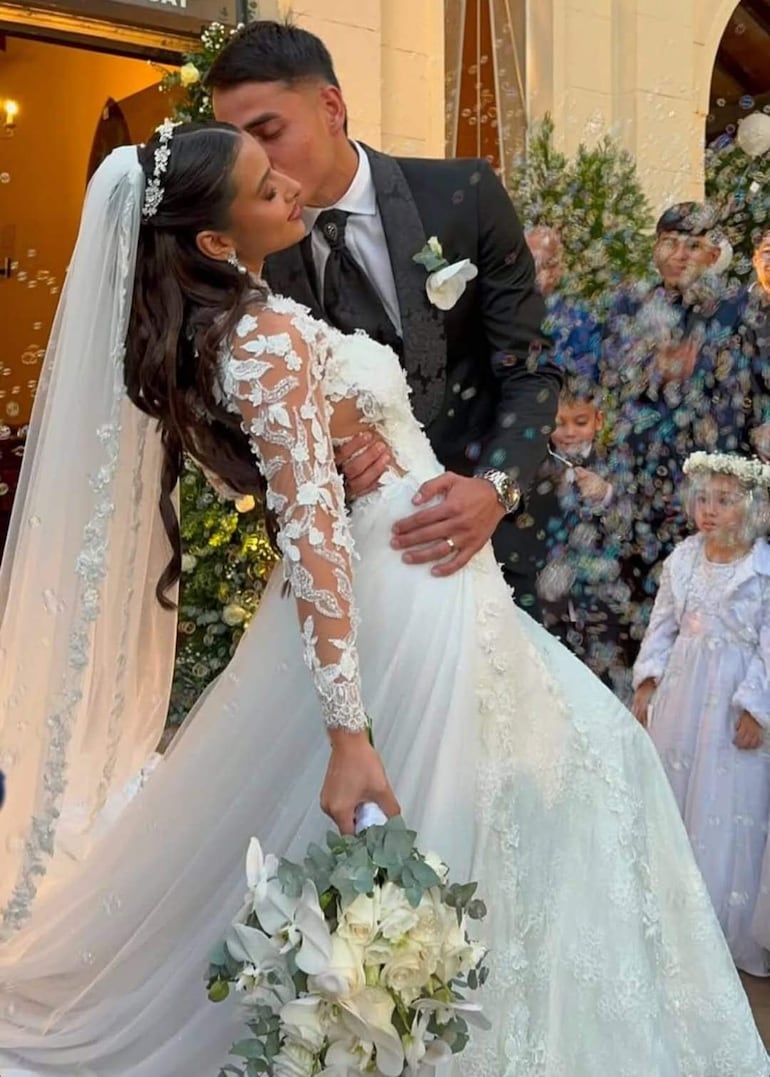 Novia en vestido blanco y novio formal, se besan en la mejilla rodeados de burbujas y niños atentos en un ambiente alegre.