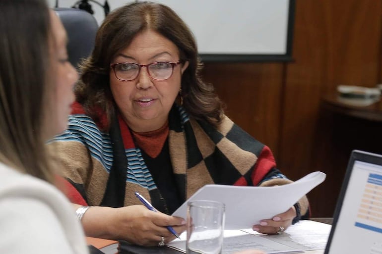 Mujer de cabello castaño con abrigo de cuadros sostiene documentos, mientras otra mujer con chaqueta clara habla en un entorno formal.