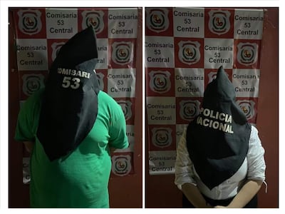 Dos personas con capuchas negras, un hombre con camiseta verde y una mujer con camisa blanca, están en una comisaría con mural visible.