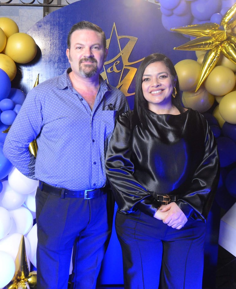 Hombre con camisa azul y pantalón oscuro junto a mujer con blusa negra y pantalón oscuro, ambos sonriendo en ambiente festivo.