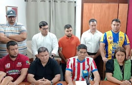 Grupo de hombres serios en conferencia; cuatro sentados con camisetas de diferentes colores, uno hablando. Ambiente interno con micrófono.