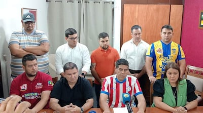 Grupo de hombres serios en conferencia; cuatro sentados con camisetas de diferentes colores, uno hablando. Ambiente interno con micrófono.