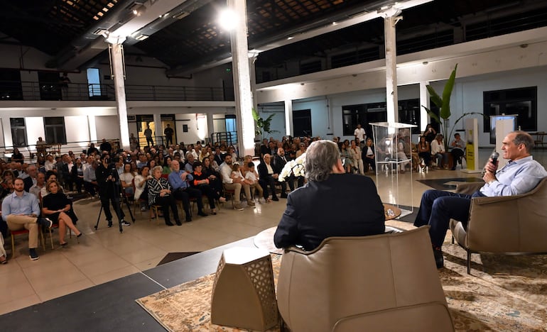 Dos hombres en sillones, uno habla con micrófono, el otro escucha; audiencia atenta en ambiente decorado con plantas.