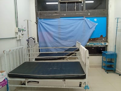 Sala de hospital con dos camas vacías y un carrito azul, luz natural y ambiente deteriorado.