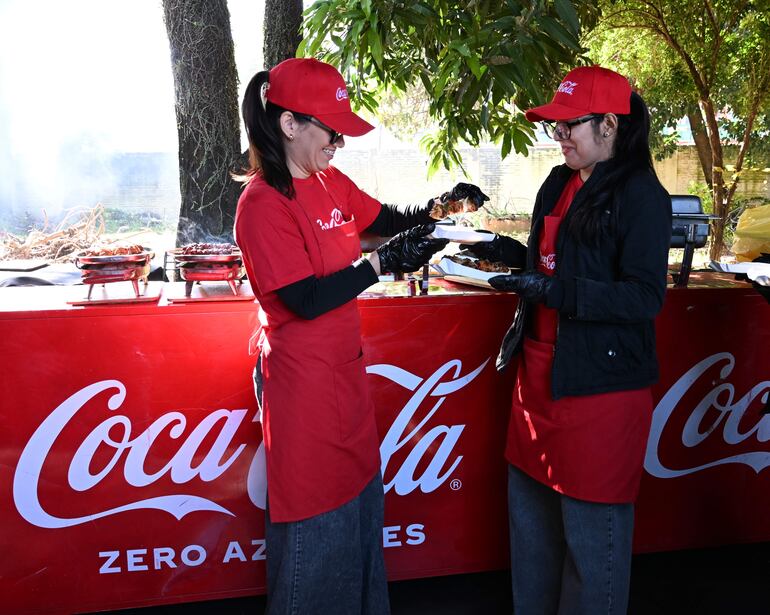 Dos mujeres con camisetas rojas y guantes negros sonríen al intercambiar un plato en un ambiente festivo.