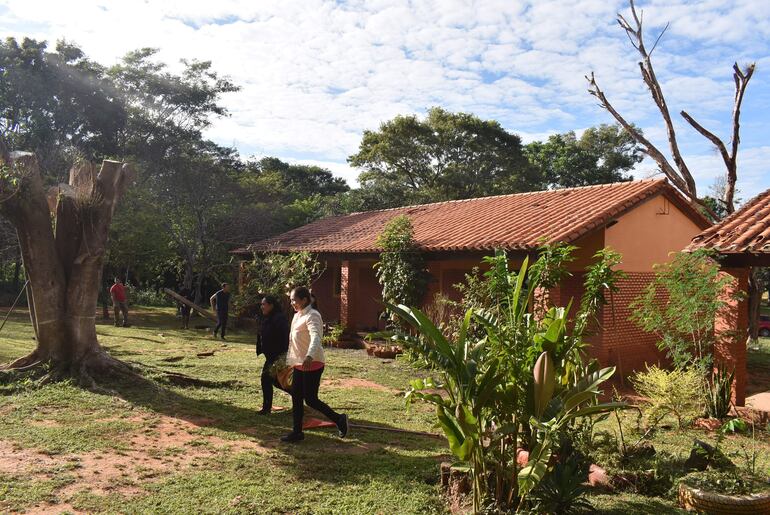 Dos mujeres conversan vestidas con chaquetas, tres hombres trabajan en el fondo rodeados de árboles y naturaleza.