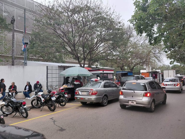 Cinco personas sentadas en una banca, dos en motocicletas, con un carrito de venta de alimentos y varios vehículos en movimiento.