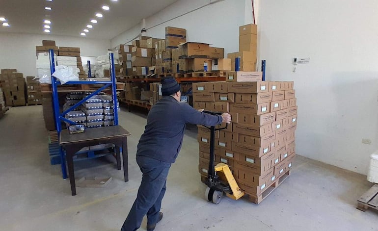 Persona de espaldas en abrigo oscuro y gorro empujando un carro cargado de cajas en un depósito iluminado.