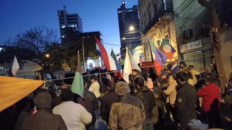 Grupo de campesinos reunidos en Asunción, sosteniendo banderas, algunos escuchan a un orador en un ambiente urbano.
