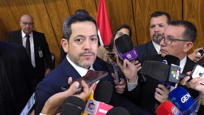 Raul Latorre Martínez vestido con traje oscuro y corbata roja, rodeado de periodistas con micrófonos en un ambiente de atención.