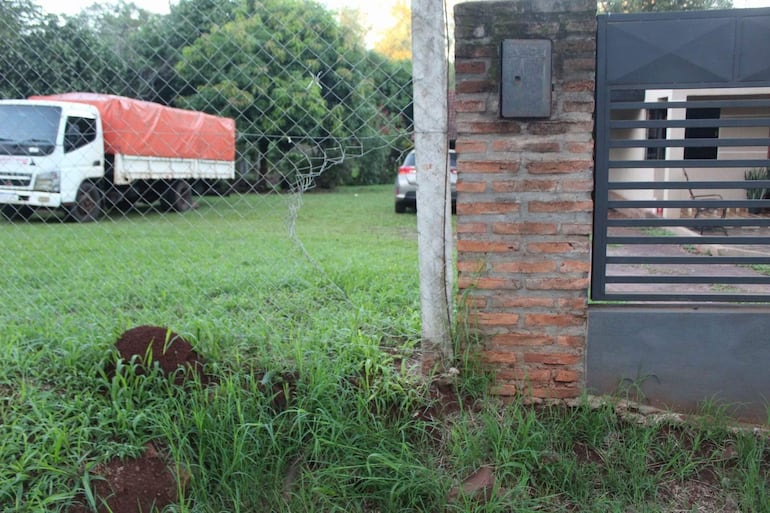 Escena exterior de residencia con portón de metal, camión blanco y coche gris estacionados en césped.