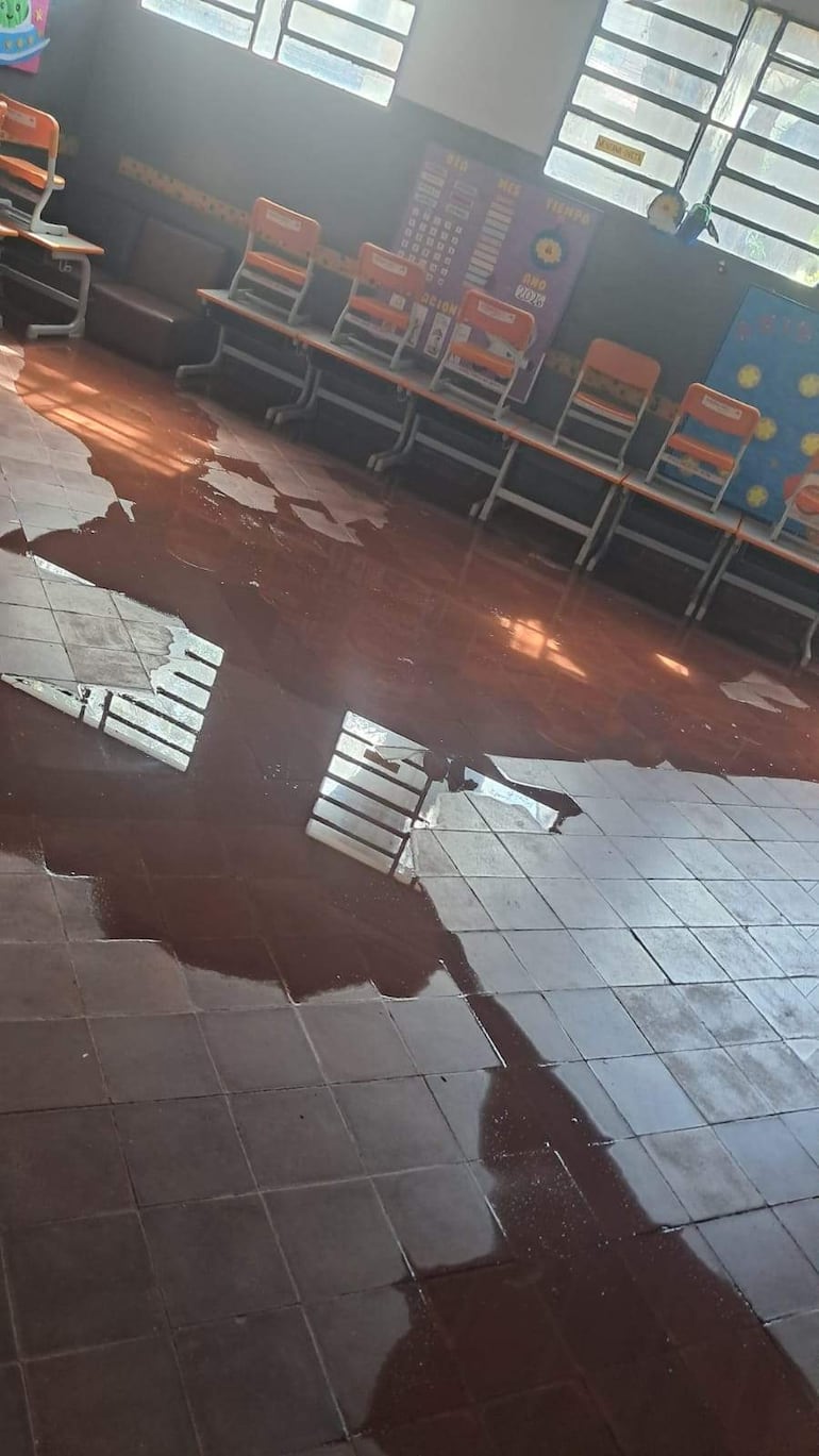 Aula vacía con suelo inundado y sillas naranjas apiladas, decoraciones educativas en la pared y luz natural entrando por las ventanas.