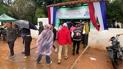 Hombres y mujeres con abrigos y paraguas se dirigen a la entrada del colegio bajo la lluvia.