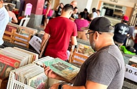Hombre con gafas y gorra revisa un disco en un mercado de vinilos lleno de personas entusiastas.