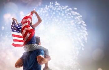 Patriotic holiday. Child sitting on shoulders of her father and holding the Flag of the USA. America celebrate 4th of July.