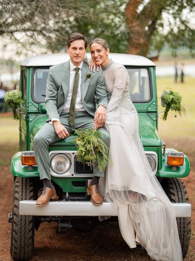 Emma Viedma luciendo un vestido blanco con detalles plateados, sonríe abrazada a Vicente Scavone en traje claro con un ramo de flores.