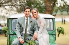 Emma Viedma luciendo un vestido blanco con detalles plateados, sonríe abrazada a Vicente Scavone en traje claro con un ramo de flores.