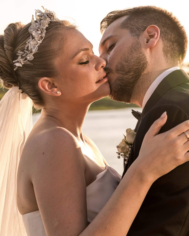 Mujer en vestido blanco y tocado junto a hombre en traje negro, casi a punto de besarse en un paisaje natural al atardecer.