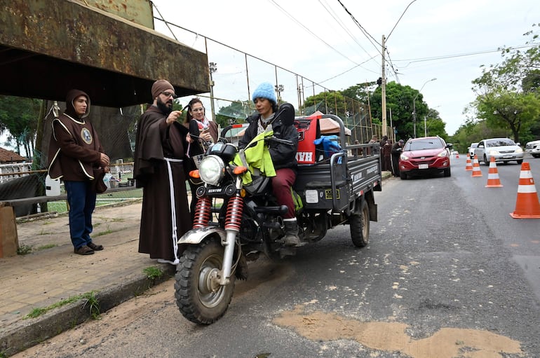 Dos hermanos capuchinos en hábito religioso, uno hablando con micrófono, público en un entorno urbano con vehículos.