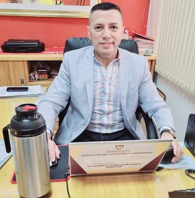 Hombre con cabello corto y blazer claro sonríe en oficina, junto a un cartel que indica su cargo como Director de Catastro.