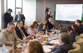 Blanca Ovelar habla con micrófono, rodeada de hombres y mujeres en una mesa con agua y comida, en un ambiente de reunión formal.
