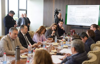 Blanca Ovelar habla con micrófono, rodeada de hombres y mujeres en una mesa con agua y comida, en un ambiente de reunión formal.