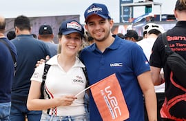 Hombre y mujer sonríen con gorra, camiseta y una bandera del WRC, rodeados de público en un evento al aire libre.
