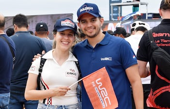 Hombre y mujer sonríen con gorra, camiseta y una bandera del WRC, rodeados de público en un evento al aire libre.