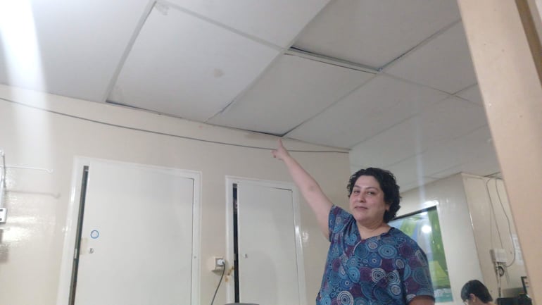 Médica de cabello rizado y blusa azul y blanca señala al techo mientras habla, en un ambiente interior sencillo.