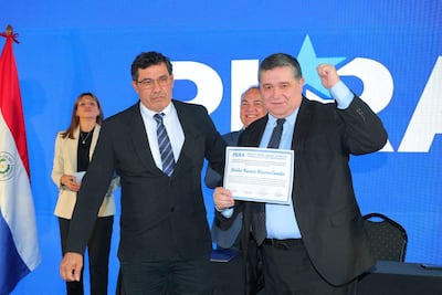 Alcides Riveros sonríe levantando un puño, mientras un hombre serio le entrega un documento en un evento del PLRA.
