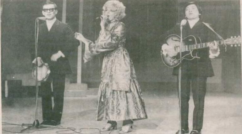Cantante en vestido llamativo al centro, guitarrista a la derecha y percusionista a la izquierda, todos sobre un escenario con iluminación básica.