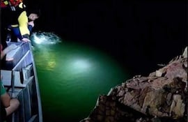 Hombres en embarcación iluminan el agua con linterna, algunos visten chalecos salvavidas. Ambiente oscuro y rocoso.