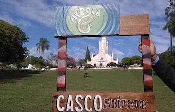 Marco de madera que dice 'CASCO ABIERTO' sostenido por una mano, rodeado de césped y árboles en un entorno al aire libre.