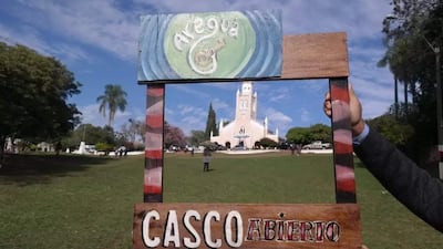 Marco de madera que dice 'CASCO ABIERTO' sostenido por una mano, rodeado de césped y árboles en un entorno al aire libre.