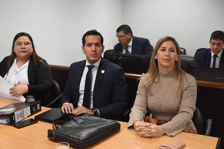 Mujer de cabello oscuro y chaqueta negra sostiene documentos; hombres en traje observan atentamente en ambiente institucional.