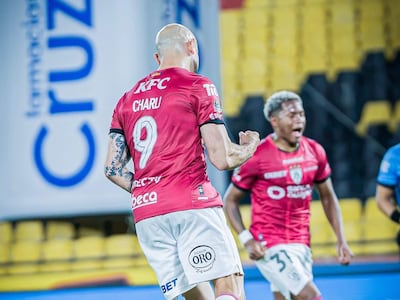El jugador CHARU, con camiseta rosa, celebra un gol levantando una mano en un estadio con gradas amarillas y negras.