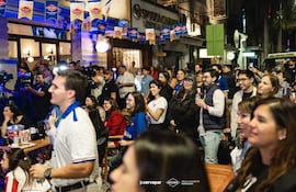 Hombres y mujeres sonrientes en un evento al aire libre, con camisetas de equipo y banderas de cerveza, creando un ambiente festivo.