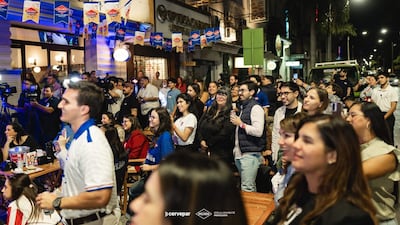 Hombres y mujeres sonrientes en un evento al aire libre, con camisetas de equipo y banderas de cerveza, creando un ambiente festivo.