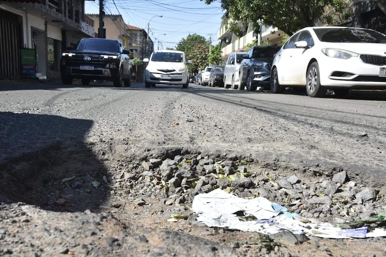 Calle urbana con bache visible, coche blanco en primer plano y vehículos en movimiento, rodeada de casas y árboles.