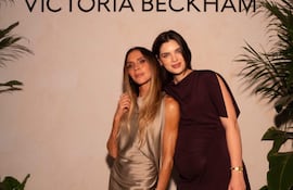 Nadia Ferreira con vestido plateado y cabello suelto, junto a Victoria Beckham, embarazada y con vestido púrpura, sonríen ante un fondo decorado.