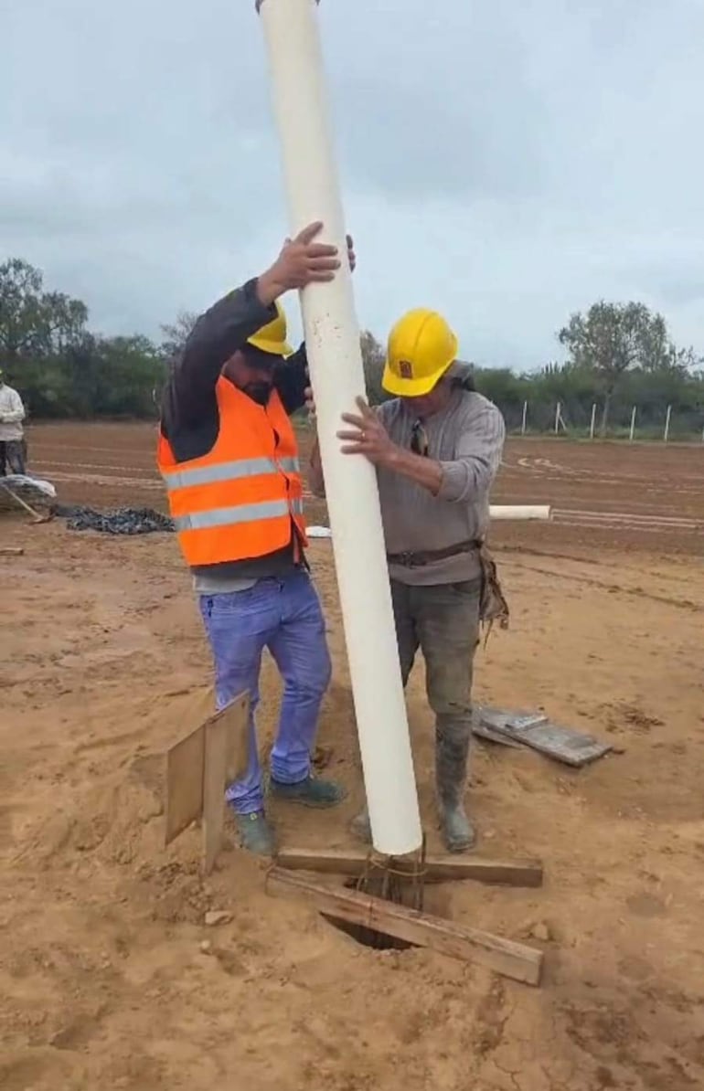 Dos trabajadores con cascos amarillos y chalecos reflectantes sosteniendo un tubo blanco en un sitio de construcción en el Chaco.