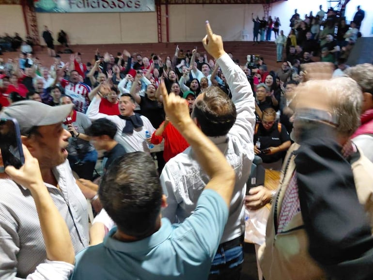 Hombres en camisetas coloridas celebran, levantando las manos con expresiones de alegría en un salón de eventos festivo.