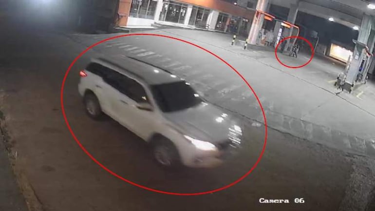 Vista de estación de servicio con SUV blanca en movimiento y luces encendidas. No hay personas claramente visibles.