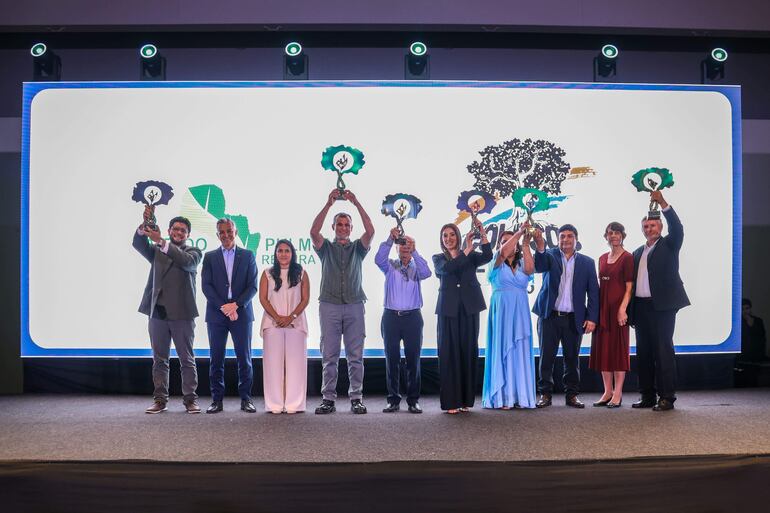Grupo de ocho personas en el escenario, todos felices y en trajes formales, sosteniendo trofeos al aire en un ambiente festivo.