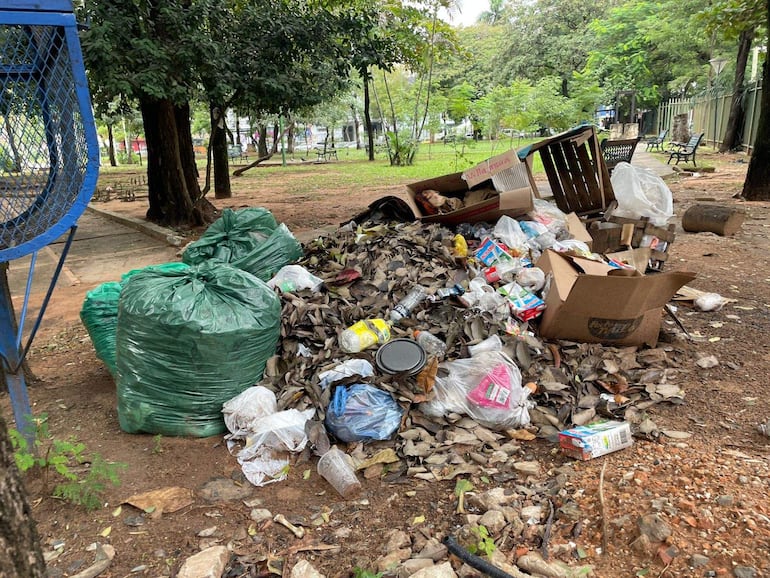 Área de parque con basura acumulada, incluyendo bolsas verdes y cartones; entorno descuidado sin personas visibles.