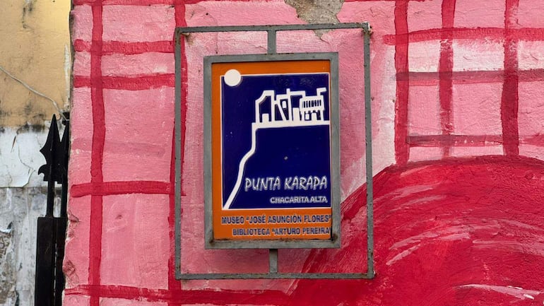 Letrero rectangular azul con detalle blanco y naranja, indicando el Museo José Asunción Flores y Biblioteca Arturo Pereira en un entorno artístico.